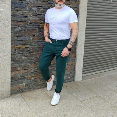 Homem com t-shirt branca, calças verde-escuras e ténis brancos encostado numa parede de pedra