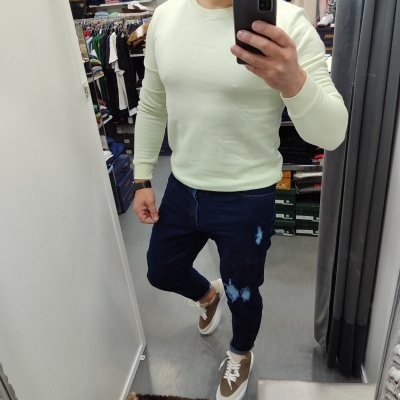Homem de sweat branco creme, jeans azul escuro com manchas claras e ténis castanhos e brancos numa loja
