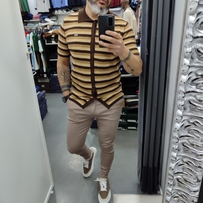 Homem a tirar selfie em espelho usando camisa polo às riscas castanhas e bege, calças justas bege e sapatilhas castanhas e brancas.