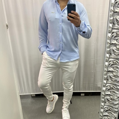 Homem com camisa azul clara, calças brancas e ténis brancos a tirar selfie num espelho.