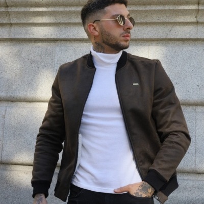 Homem com casaco castanho e camiseta branca frente a parede cinzenta de pedra