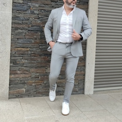 Homem com fato cinzento claro e sapatilhas brancas junto a parede de pedra e porta metálica
