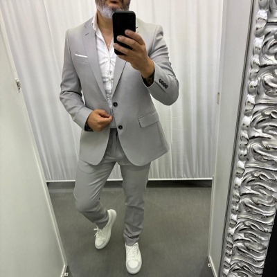 Homem a vestir fato cinza claro com camisa branca e ténis brancos em frente a espelho