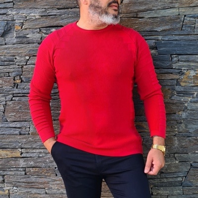 Homem com camisola vermelha de malha e calças pretas junto a parede de pedra
