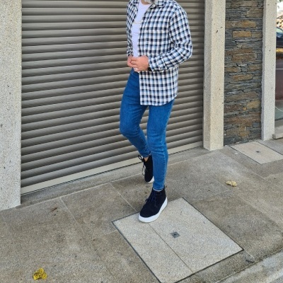 Homem com camisa xadrez, jeans azuis e sapatilhas pretas junto a parede e porta metálica