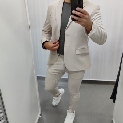 Homem a vestir fato bege claro numa cabine de provador com parede branca e chão cinzento