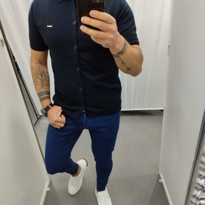 Homem vestindo camisa preta de manga curta, calças azul escuro e sapatilhas brancas num provador.