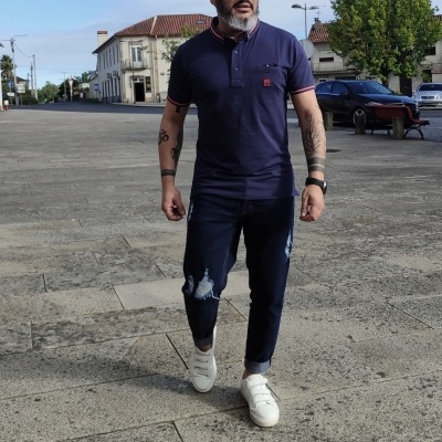 Homem com camisa polo azul escura, calças de ganga rasgadas e sapatilhas brancas a caminhar num passeio de pedra.