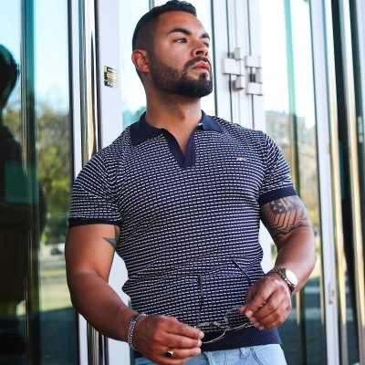 Homem a usar polo azul e branco com calças jeans, junto a parede de vidro refletivo