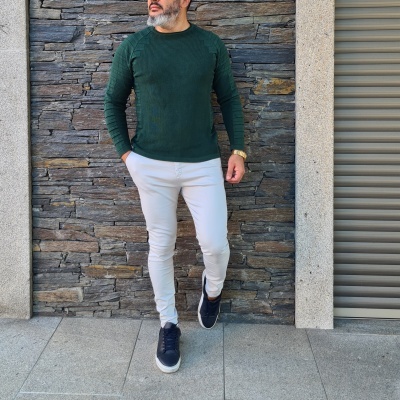 Homem com camisola verde, calças brancas e ténis pretos junto a parede de pedra