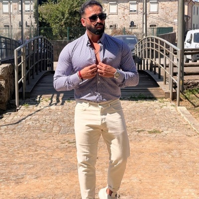 Homem com camisa azul e branca, calças bege e ténis brancos a andar numa rua com calçada e ponte ao fundo