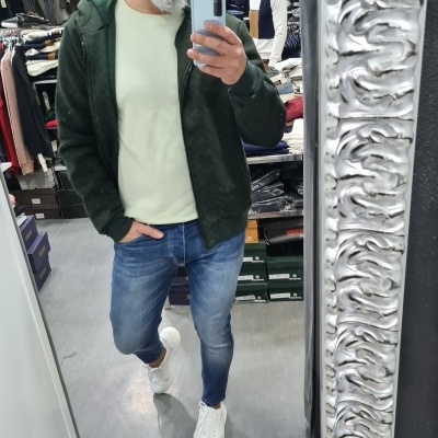 Homem com casaco verde escuro, T-shirt clara, jeans azuis e ténis brancos numa loja