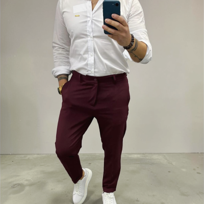 Homem com camisa branca, calças bordeaux e sapatilhas brancas, a tirar selfie num ambiente minimalista.