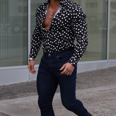 Homem com camisa preta de bolinhas brancas, calças azuis escuras e botas pretas num passeio urbano