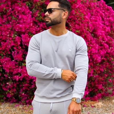 Homem de fato de treino cinza claro com riscas brancas, óculos escuros e relógio dourado diante de flores cor-de-rosa.