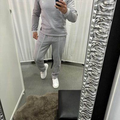 Homem a usar conjunto desportivo cinzento claro com ténis brancos em frente a um espelho prateado texturizado.