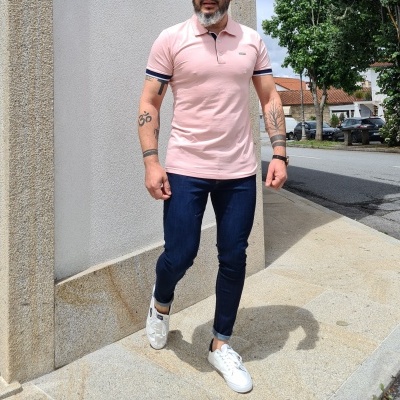 Homem com polo rosa e calças jeans azul escuro junto a um muro e rua com árvores