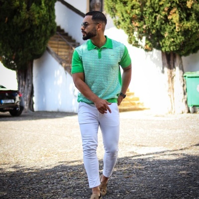 Homem com camisa polo verde e calças brancas ao ar livre com árvores e escadas ao fundo