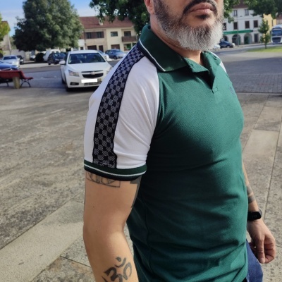 Homem a usar polo verde escuro com mangas brancas e faixa estampada preta e branca, em ambiente urbano