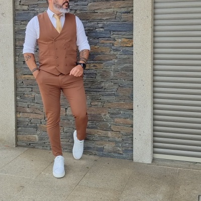 Homem com colete castanho, calças, camisa branca, gravata amarela e ténis brancos junto a parede de pedra