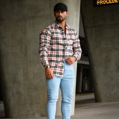 Homem com camisa xadrez, calças azuis claras e boné preto em espaço urbano com pilares de concreto