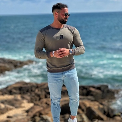 Homem vestindo camisola verde e calças jeans junto a uma costa rochosa com o mar ao fundo