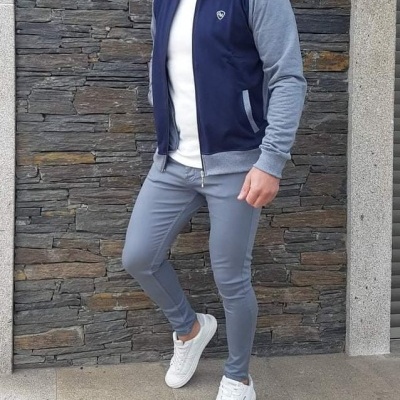 Homem com casaco azul e cinza, calças cinza e sapatilhas brancas junto a parede de pedra