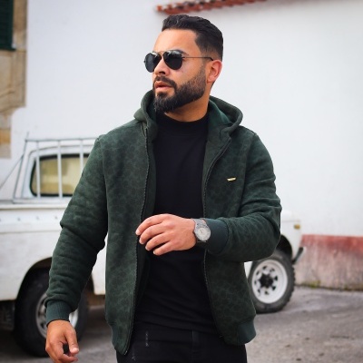 Homem com casaco verde escuro com capuz e padrão leve, usando gafas de sol e relógio prateado