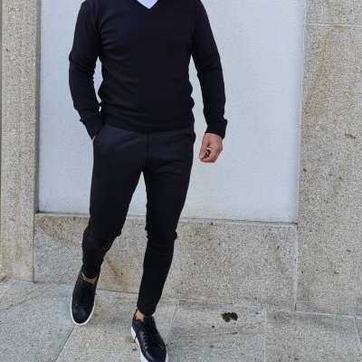 Homem com camisola preta, calças pretas e sapatos pretos junto a parede de pedra
