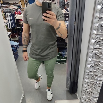 Homem de t-shirt verde-oliva, calças verdes claras e ténis brancos a tirar selfie num espelho numa loja.