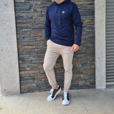 Homem com sweatshirt azul e calças bege junto a parede de pedra cinzenta