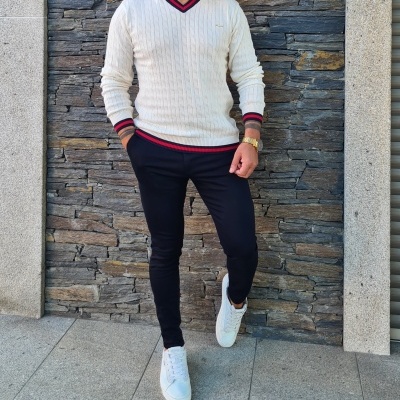 Homem com camisola branca, calças pretas e sapatilhas brancas em frente a parede de pedra.