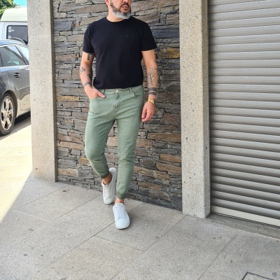 Homem com t-shirt preta, calças verdes e sapatos brancos junto a parede de pedra