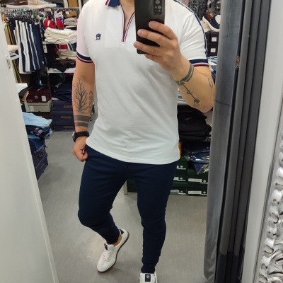 Homem a tirar selfie num espelho vestindo camisa polo branca com detalhes azuis e vermelhos e calças azuis escuras