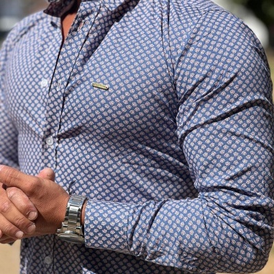 Camisa azul claro com padrão repetido em branco e rosa, botão e logotipo no peito, pulseira metálica no pulso