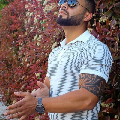 Homem com camisa pólo branca de riscas pretas e óculos escuros junto a sebe vermelha