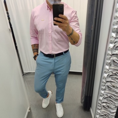 Homem vestido com camisa rosa, calças azuis e ténis brancos a tirar selfie em espelho