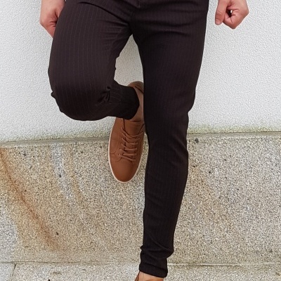 Homem vestido com calças pretas risca de giz, cinto castanho e sapatos castanhos, em fundo de parede branca e chão de cimento.