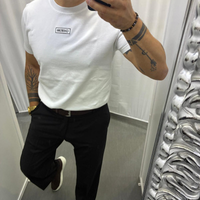 Homem de branco com t-shirt, calças pretas e pulseira a tirar selfie num espelho.