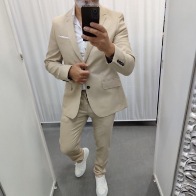 Homem num fato bege, camisa branca e sapatilhas brancas num provador com cortinas brancas