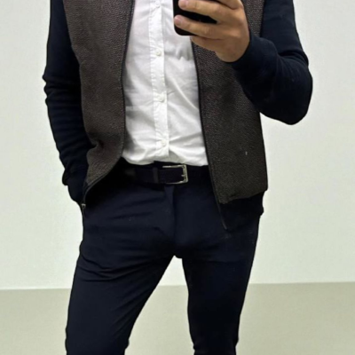 Homem de camisa branca, casaco castanho e preto, calças e cinto pretos, sapatilhas brancas, a tirar selfie com smartphone preto.