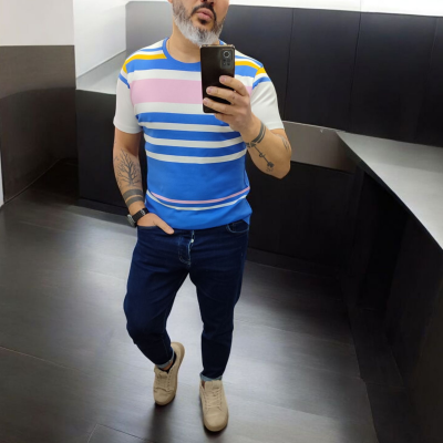 Homem a usar camisola de riscas coloridas, calças jeans e sapatilhas enquanto tira uma selfie numa casa de banho.