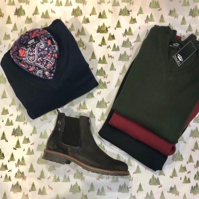 Três camisolas dobradas e uma bota de couro marrom sobre tecido branco com padrão de árvores verdes