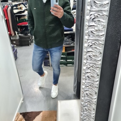 Homem com casaco verde e calças de ganga a tirar selfie num espelho com moldura prateada
