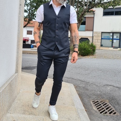 Homem com colete cinza escuro, camisa branca de manga curta e calças cinza escuro a caminhar na calçada