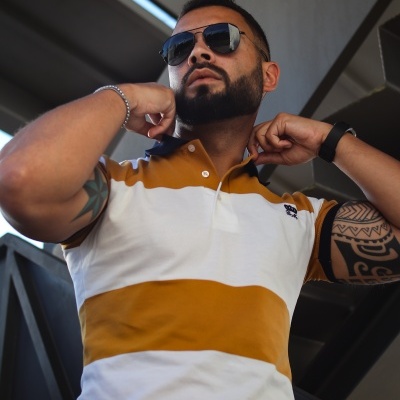 Homem com polo branco e amarelo, óculos escuros e tatuagens numa estrutura metálica industrial