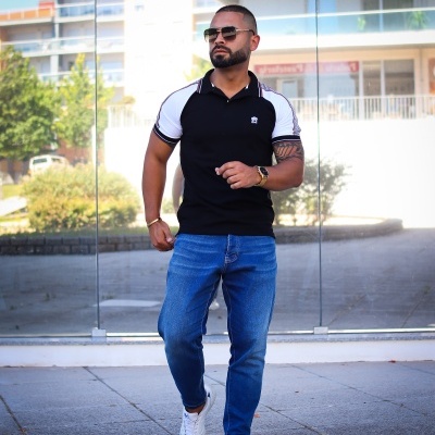 Homem com t-shirt polo preta com mangas brancas e calças de ganga azuis em ambiente urbano