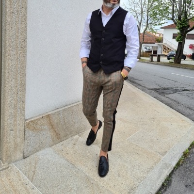 Homem com colete preto e calças xadrez ao lado de uma parede branca na rua