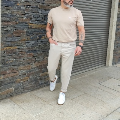 Homem com roupa casual bege e ténis brancos numa rua com parede de pedra e porta metálica.