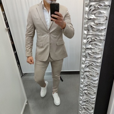 Homem com fato bege claro, camisa branca e sapatilhas brancas em frente a espelho prateado